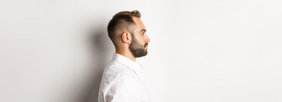 Close-up profile shot of handsome bearded man looking left, standing against white background.