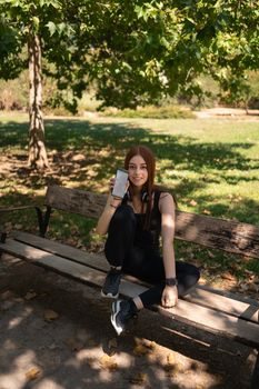 Young slim female athlete in black sportswear and headphones showing mobile phone with empty screen in park while sitting on bench
