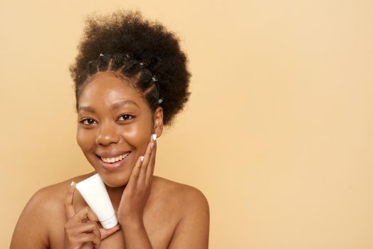 Lovely african american woman holding moisturizer or mask on beige background with copy space. Skin care, anti aging.