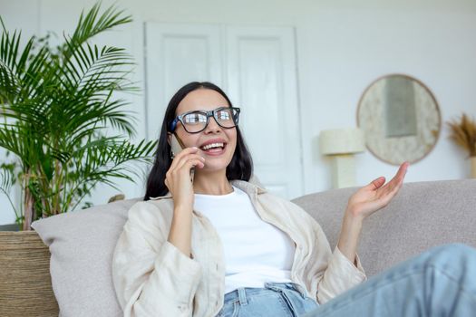 Happy young beautiful woman in glasses sitting on the sofa at home and talking on the phone with friends and family. Gesturing with hands. Quarantine, remote online communication.