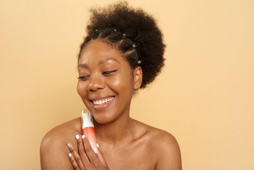 Dark-skinned woman with a wide smile, holds a moisturizer near her face, enjoys the softness of the skin, isolated on a beige background. Facial treatments.