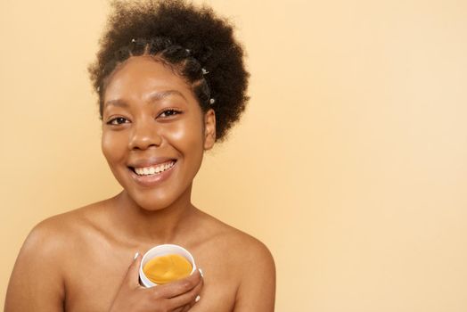 Charming cheerful curly african american woman holds cosmetic patches under her eyes, looks joyfully at the camera, poses on a beige background, copy space for your advertisement.