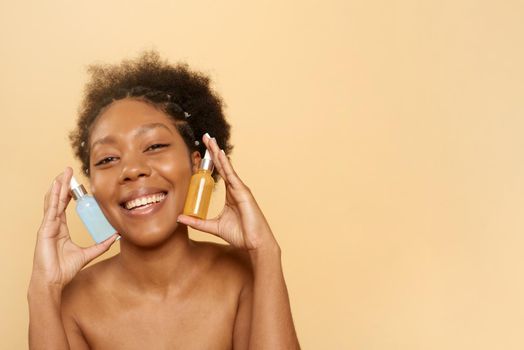 Beautiful african american woman holding bottles of hyaluronic acid for facial skin care and vitamin c serum. Black lady holding glass jars isolated on beige background with copy space and looking at camera.