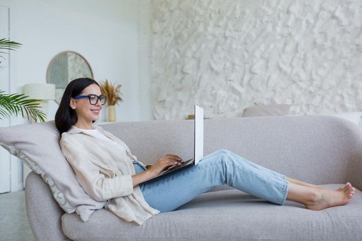 Online work. Young business woman in glasses, freelancer working at home on laptop. Lying comfortably on the couch in jeans and a shirt, typing