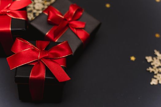 Black gift boxes with a red ribbon on a gold confetti background. Elegant Christmas background with modern golden Christmas decorations, balls, gift boxes, confetti stars on dark black background. Flat design, top view, copy space