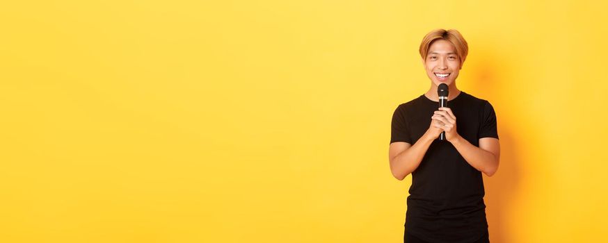 Portrait of charismatic smiling asian man holding microphone and singing karaoke, performing song, standing yellow background.