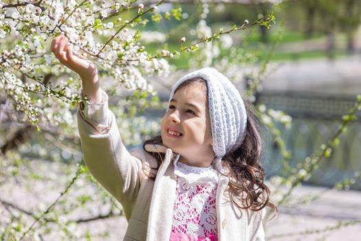 a little girl in a white coat, holding a branch of a tree blooming with white flowers, in the spring in the park in sunny day . Close up