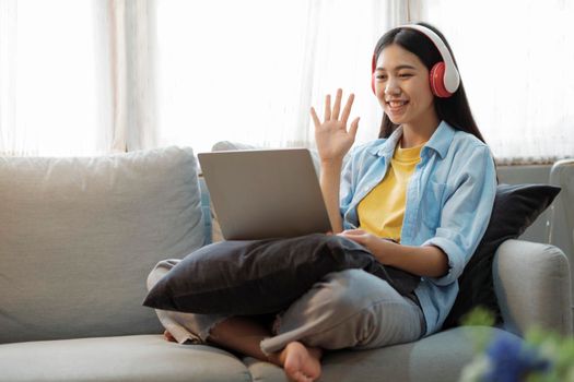 Young asian woman smiling, listening to music, and online meeting using laptop while sitting on couch at home with enjoyment. Online connection and online learning concept.