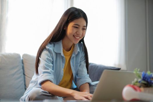 Young asian woman smiling and using laptop to study or work while sitting on couch at home. Women online learning and searching for information using laptop while sitting comfortably on couch in the bright living room. Working, studying on desk at home. online learning