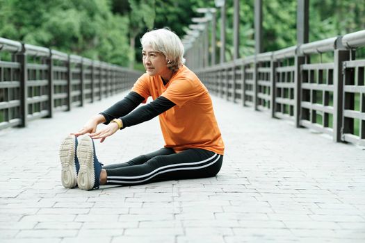 Athletic Senior woman jogger warming up by stretching two legs and upper body before running in park, health concept.