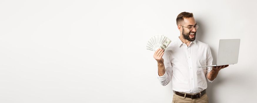 Business and e-commerce. Satisfied businessman doing job on laptop and holding money, smiling happy, standing over white background.