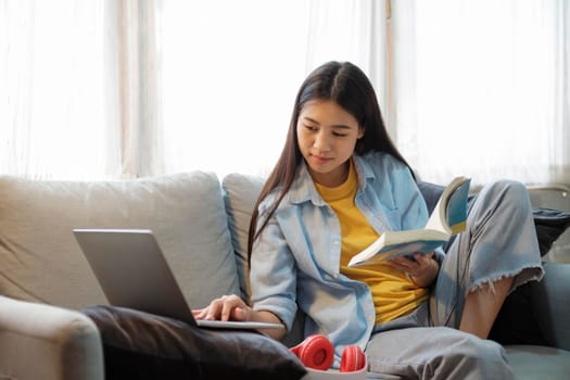 Young asian woman surfing the internet using laptop and holding book while sitting on couch at home. Young asian women studying online at home. Online connection and online learning concept.