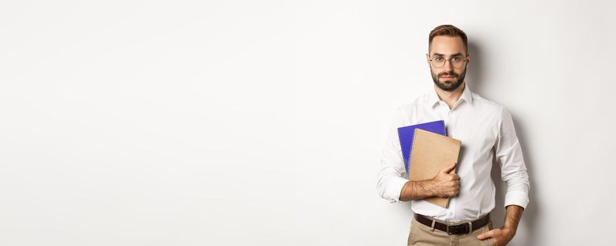 Handsome male employee holding notebooks, looking confident, standing over white background.