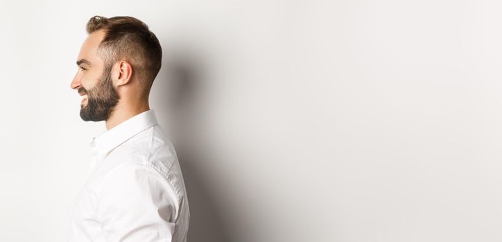 Close-up profile shot of handsome bearded man looking left and smiling, standing against white background.