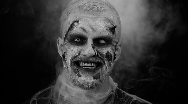 Creepy man with bloody scars face, Halloween stylish zombie make-up. Scary wounded undead guy making faces, looks at camera and smiles terribly. Voodoo rituals. Thematic party. Sinister beast, monster