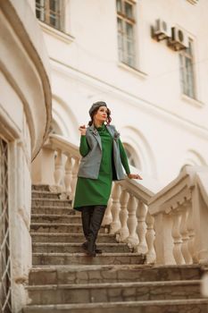 Outdoor fashion portrait of an elegant fashionable brunette model in a beret, green dress and a gray waistcoat posing at sunset in a european city in autumn