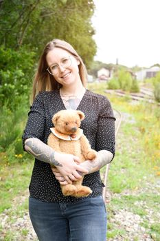 a young caucasian girl with glasses and tattoos smiles, a brown teddy bear sits in a copper pot against the background of an abandoned railway and old rails. High quality photo