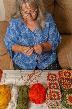 older woman with white hair and glasses crocheting on the sofa at home