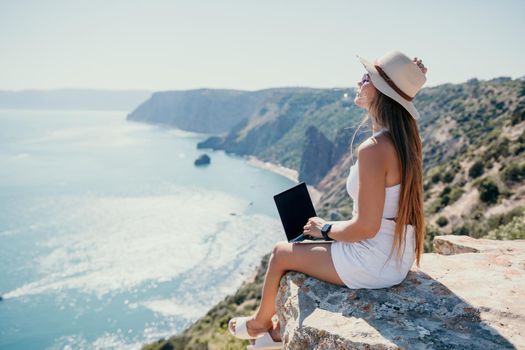 Successful business woman in yellow hat working on laptop by the sea. Pretty lady typing on computer at summer day outdoors. Freelance, travel and holidays concept.
