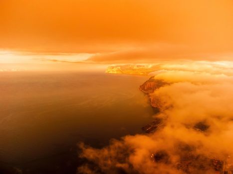 A red burning sunset with the silhouette of a cliff over the sea. Abstract nature summer or spring ocean sea background. Small waves on golden warm water surface with bokeh lights from sun.
