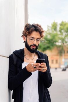 portrait of a stylish young man with beard leaning in a white wall typing on mobile phone, concept of urban lifestyle and technology of communication, copy space for text