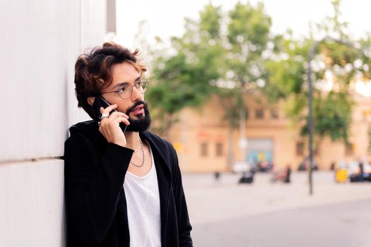 portrait of a stylish young man with beard leaning in a white wall talking on mobile phone, concept of urban lifestyle and technology of communication, copy space for text