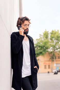 stylish young man with beard leaning in a white wall talking on mobile phone, concept of urban lifestyle and technology of communication, copy space for text