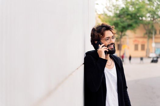 smiling young man with beard leaning in a white wall talking happy on mobile phone, concept of urban lifestyle and technology of communication, copy space for text