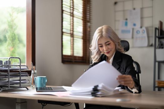 Young asian businesswoman working with laptop, financial charts and graphs reviewing data in paper documents making money work at office desk.