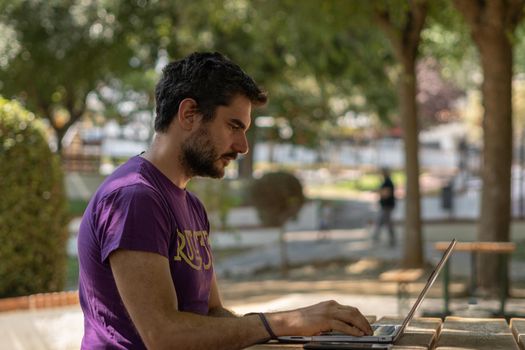 young man working at his computer on a wooden table in the park on a sunny day