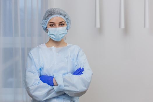 Portrait of a young female doctor in a mask, surgical gown and hat, standing with arms crossed, looking at camera