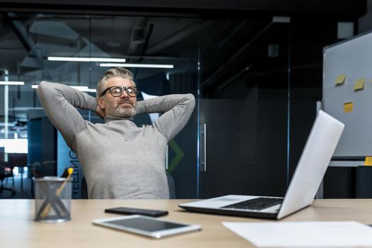 Senior gray haired man resting in office on chair with hands behind head, businessman boss working using laptop, mature investor completed project, dreaming.