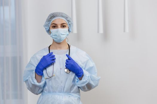 Portrait of young female doctor in mask, surgical gown and hat, standing holding stethoscope, looking at camera