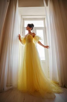 A woman's silhouette in a golden luxurious dress against the background of a window holds a curtain with her hands. Elegant lady in a yellow long silk dress with bare back, back view