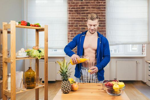 Young athletic fitness handsome man pours a glass of fresh orange juice from glass in kitchen at home, looking at camera, smiling