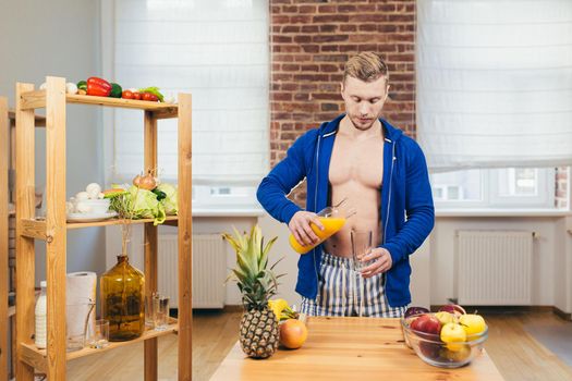 Young athletic fitness handsome man pours a glass of fresh orange juice from glass in kitchen at home, looking at camera, smiling