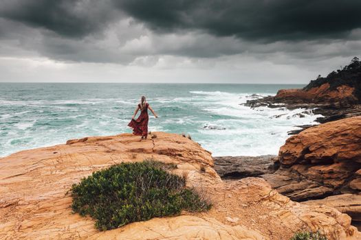 Brave female with long flowing dress, stands alone on a rock by the ocean and faces the dark storm
