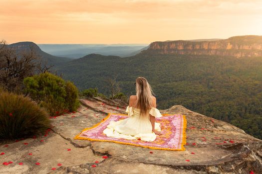 Woman wearing soft pale yellow dress sits on a pretty woven rug on cliff top with magnificent mountain views