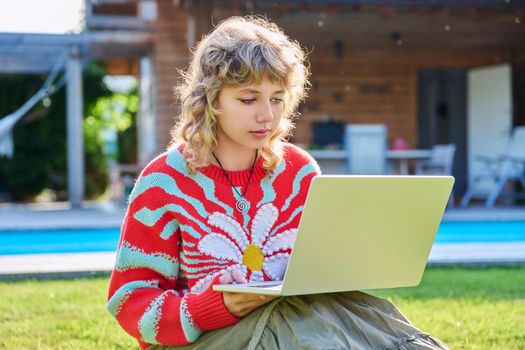 Teenage girl 17 years old sitting on lawn in backyard, using laptop for studying leisure. Adolescence, students, high school, technology, lifestyle, youth concept