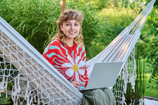 Portrait of teenage girl with laptop outdoor, smiling cute female teenager posing looking at camera, sitting on backyard lawn. Beautiful girl 16, 17 years old with curly hair, high school student