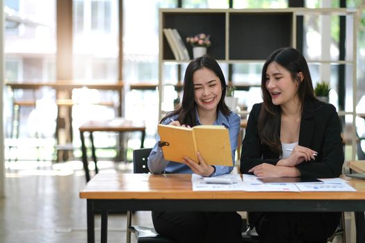 Two pretty young Asian businesswoman sitting at desk with laptop doing paperwork together discussing project financial report. Corporate business collaboration concept..