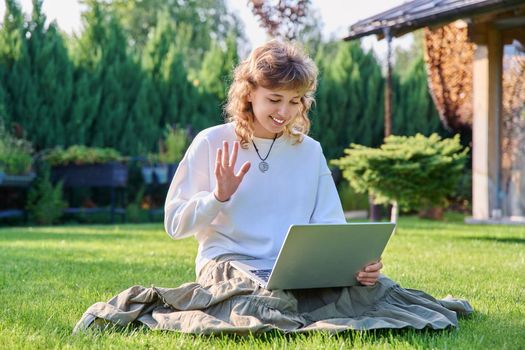 Teen girl using laptop for video communication, waving hand looking at screen, sitting on lawn green grass in backyard. Technology, chat conference video call, lifestyle, adolescence