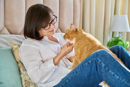 Middle aged woman talking with ginger pet cat, home interior background. Friendship, love, care, leisure, lifestyle, animals people house concept