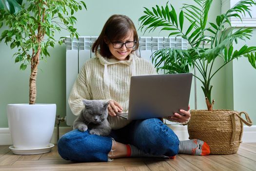 Middle-aged woman in warm clothes with cat using laptop. Female warming at home near heating radiator, with pet in her arms. Cold season winter autumn, leisure lifestyle work freelance, people animals