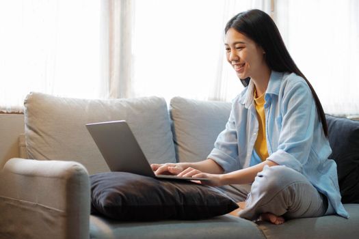 Young asian woman smiling and using laptop to study or work while sitting on couch at home. Women online learning and searching for information using laptop while sitting comfortably on couch in the bright living room. At home. online learning