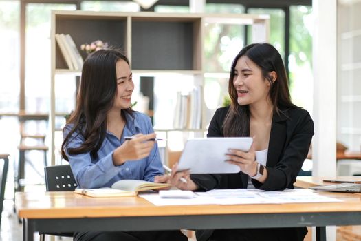 Two pretty young Asian businesswoman sitting at desk with laptop doing paperwork together discussing project financial report. Corporate business collaboration concept..