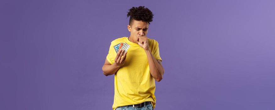 Health, influenza, covid-19 concept. Portrait of ill young man caught cold, have severe caugh, holding pills, showing drugs bought in drugstore, standing purple background feeling bad.