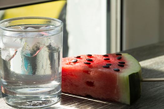 A triangular piece of sweet watermelon on an ice cream shelf and a cold drink with ice in a cafe on a wooden surface.,,