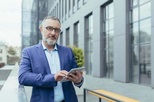 A handsome senior gray-haired man in a suit and glasses holds a tablet in his hands and reads the news. Standing near a modern office center, looking at the camera.