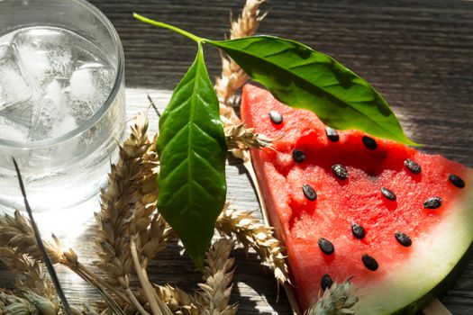 A triangular piece of sweet watermelon on an ice cream shelf, a cold drink with ice and ripe wheat ears on a wooden surface.,,